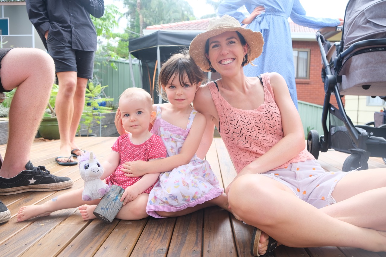 Ali Gerritsen, smiling mum with two young girls sitting on timber deck