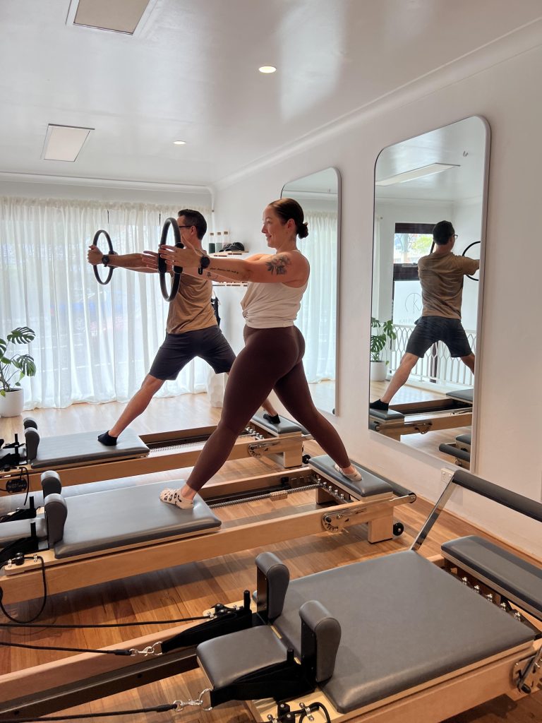 A man and a woman performing Pilates exercises using reformer machines in a bright, airy studio with wooden floors and large mirrors.