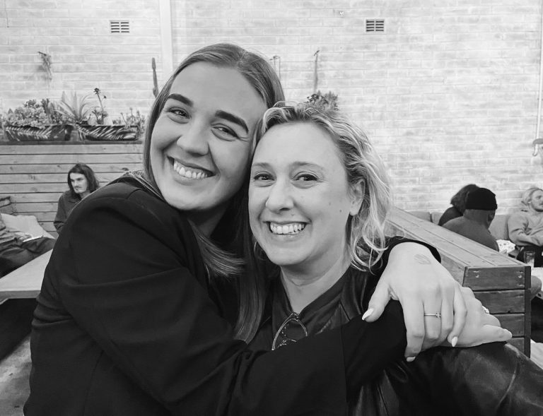 A black-and-white photo of two women smiling and embracing each other. They are both looking at the camera with joy. The background shows a brick wall and people sitting on benches.