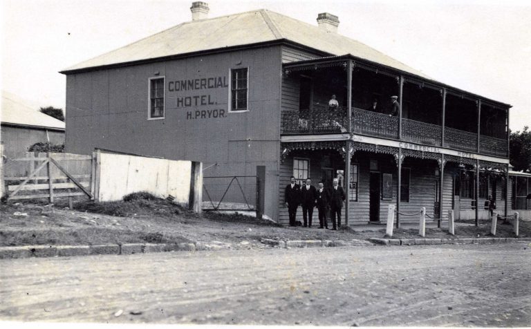 A historic black-and-white photo of the Commercial Hotel in Jamberoo, featuring a two-story building with ornate ironwork on the balcony and several men standing outside.