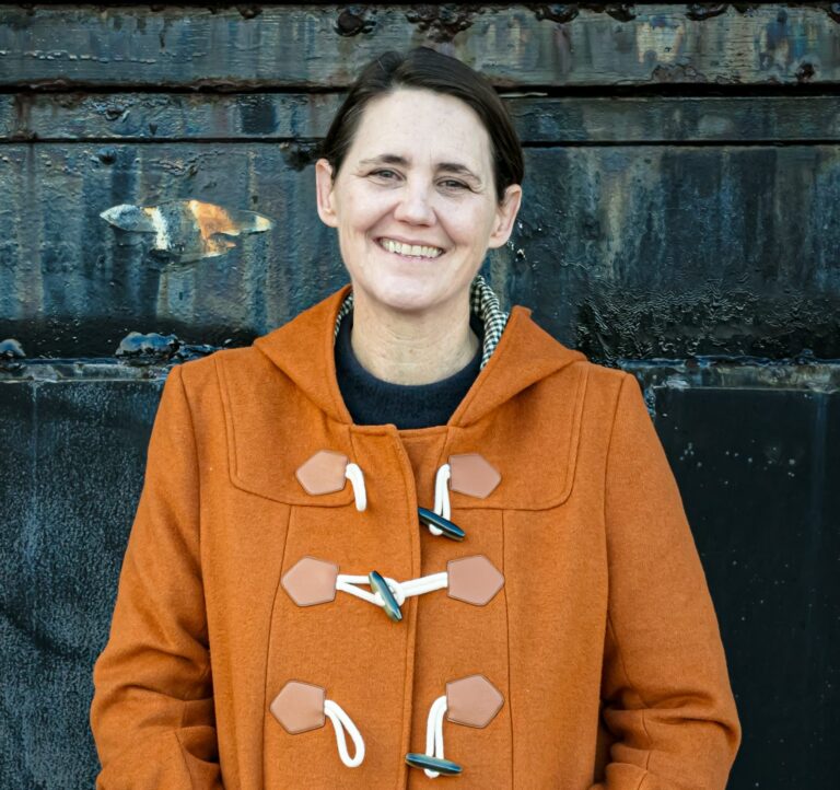 A woman with short dark hair wearing an orange coat smiles at the camera, standing against a rustic dark background with her hands in her pockets.