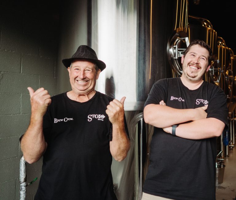 Two men, one older and one younger, smiling and giving thumbs up in front of large stainless steel beer vats at Stoic Brewing.