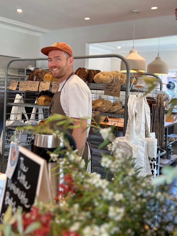 A man wearing a white shirt and an apron, smiling and standing in front of shelves filled with bread in a bakery. There are flowers in the foreground.